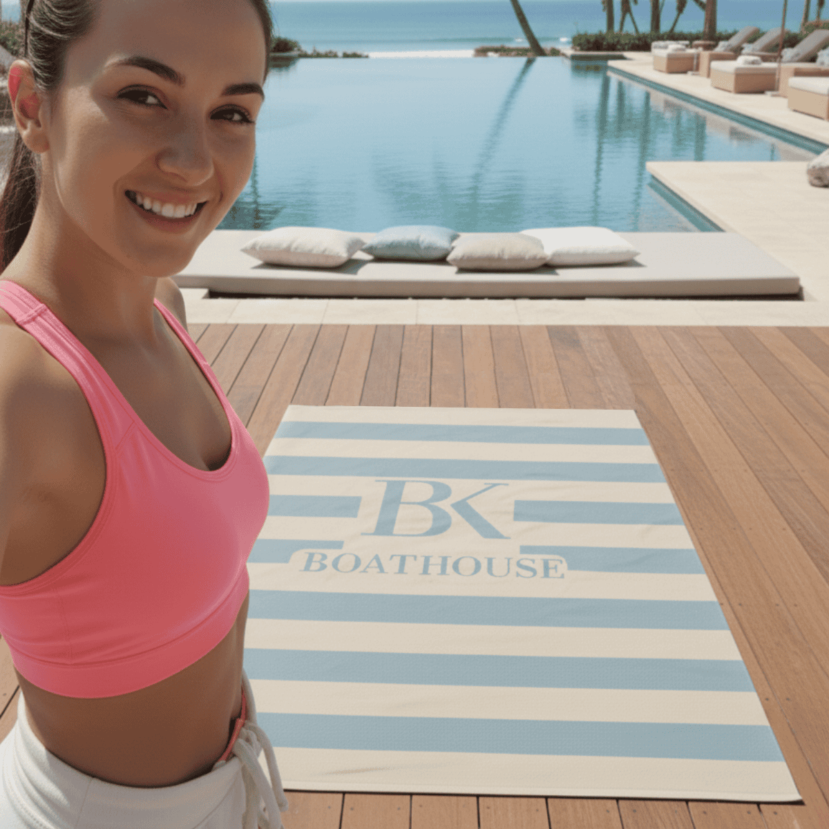 Woman in pink sports bra standing next to a striped towel with 'BK Boathouse' logo by a pool.